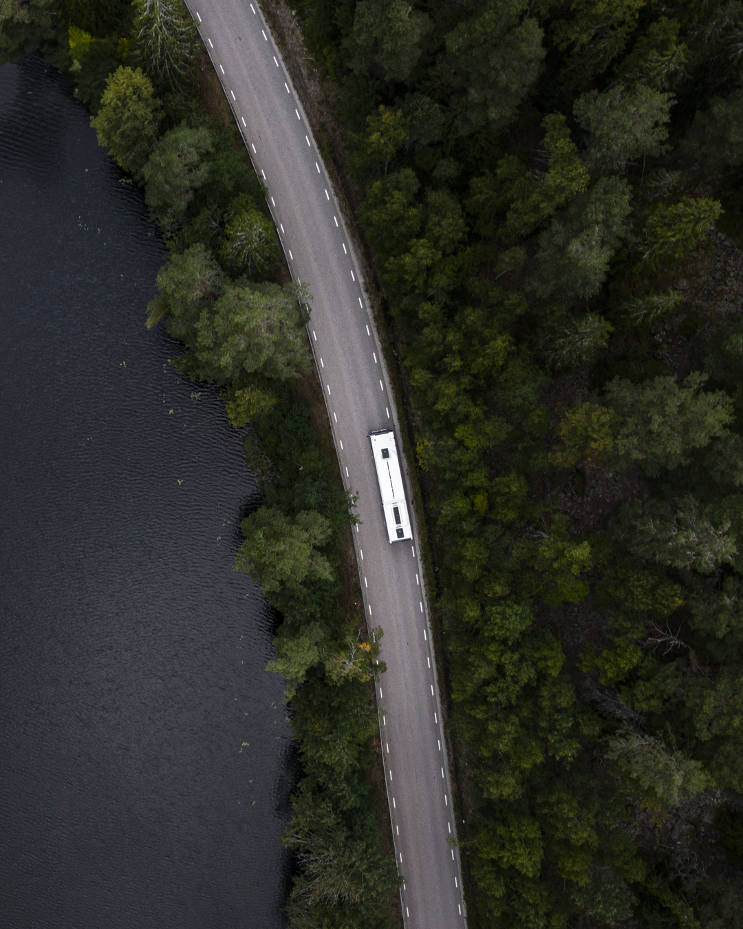 Buss fotad uppifrån som kör på en väg i en skog, bredvid vatten.