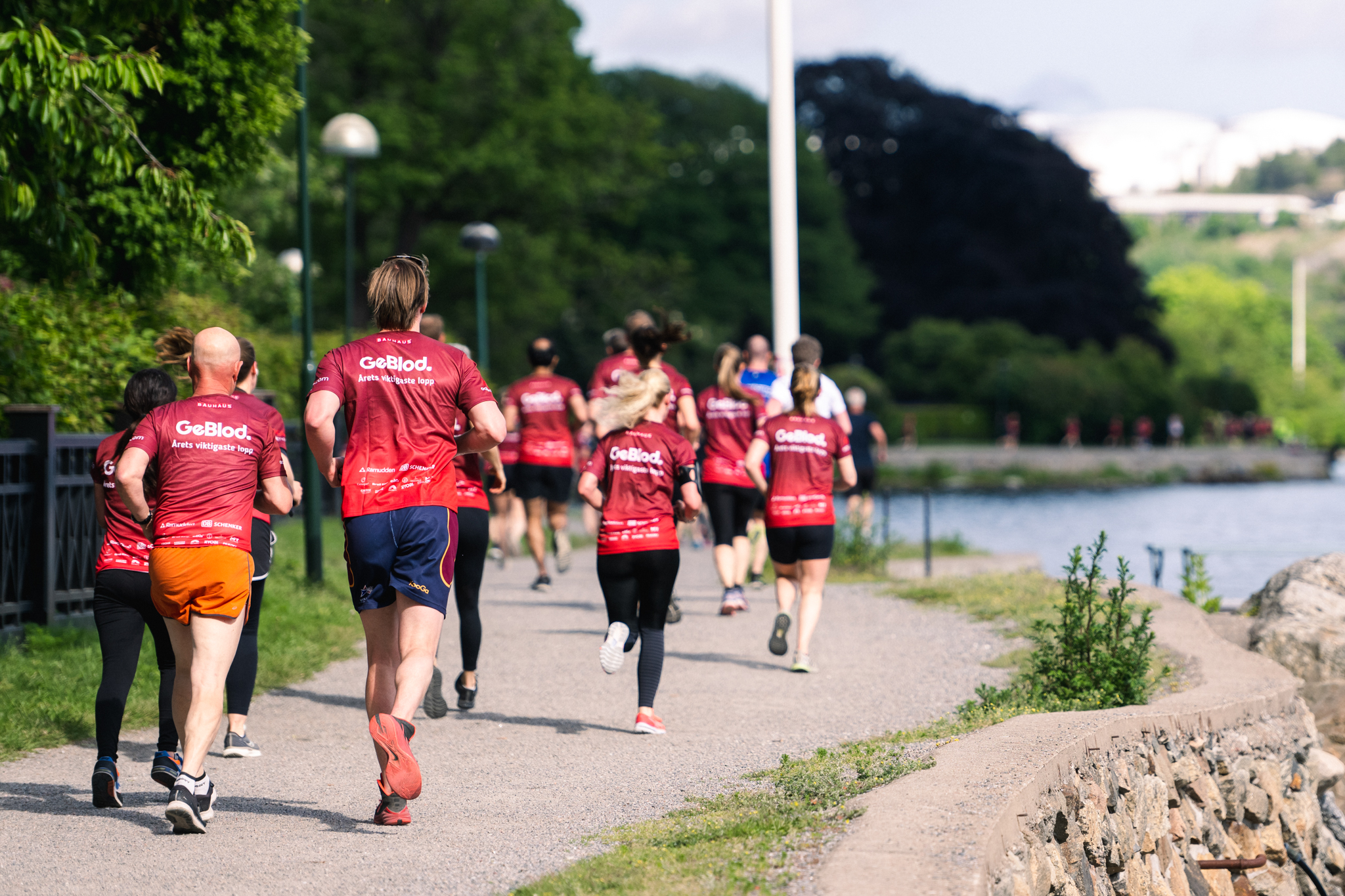 Människor i likadana t-shirts som springer längs med vatten, med träd och buskar runt omkring.